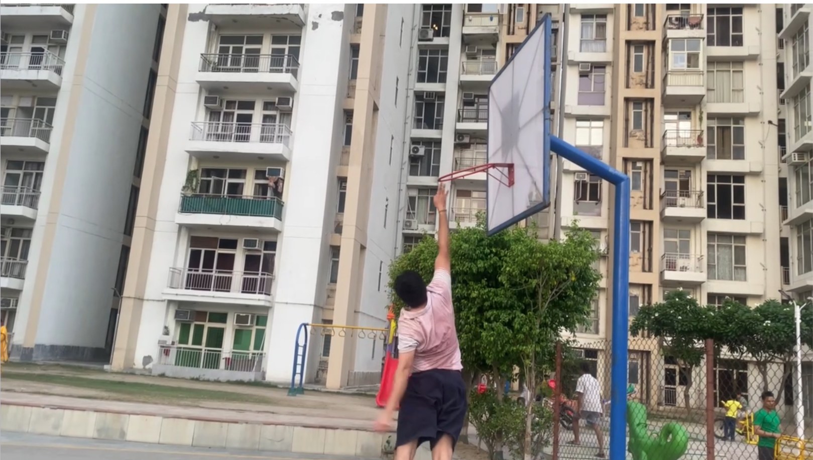 Kshetez from behind, mid jump, reaching for a basketball rim on an outdoor court with apartment towers behind him